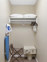 A small, neatly organized room with a bed, an ironing board, a chair, and a desk with a computer.