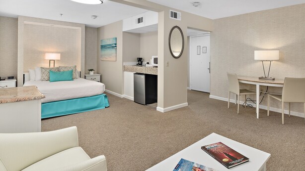 A hotel room with a bed, a desk, a chair, a small kitchenette, and a bathroom. - Brighton Suites Hotel (Rehoboth Beach)