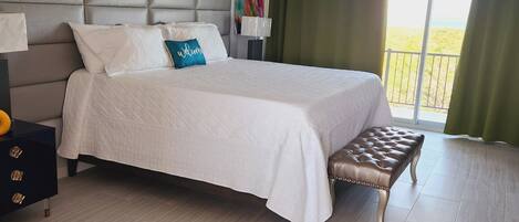 A hotel room with a large bed, a nightstand, a bench, a colorful painting, and a view of the outdoors.