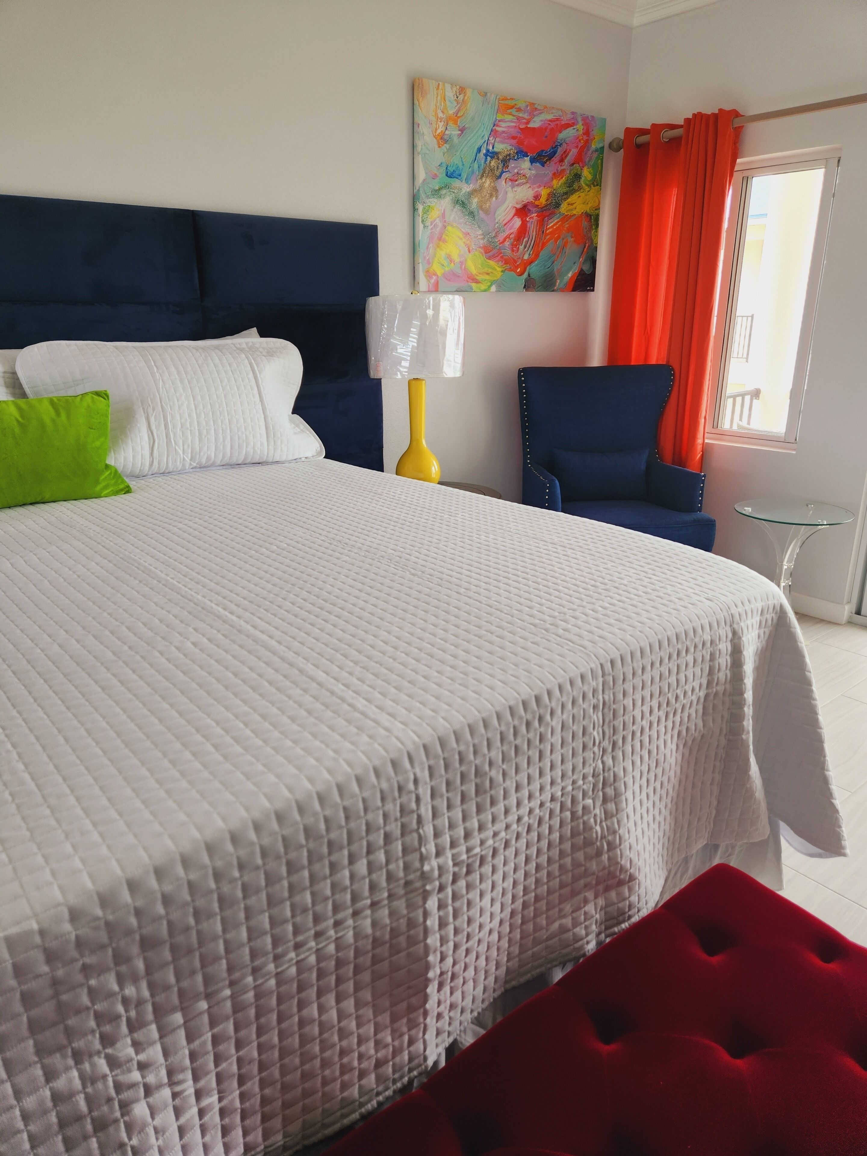 A hotel room with a large bed, a blue armchair, a small table, and a colorful abstract painting on the wall.