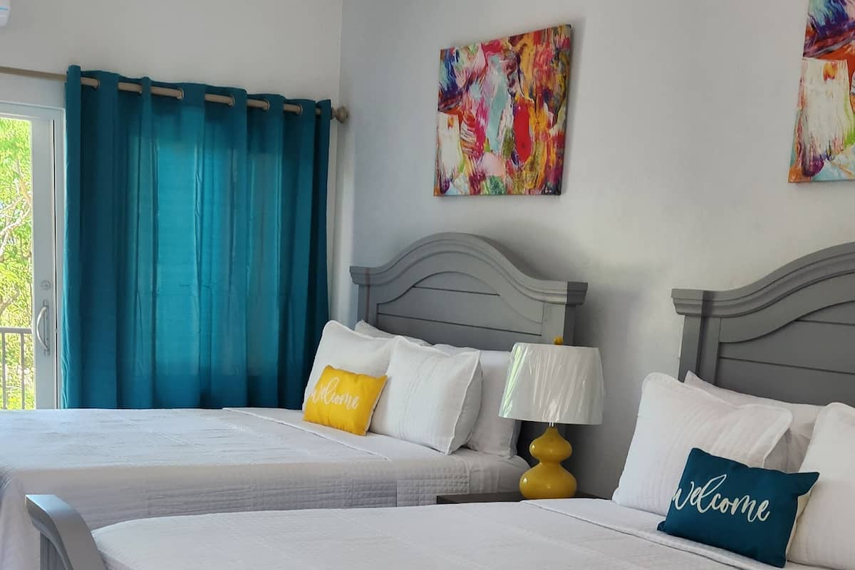 Une chambre d’hôtel avec deux lits, chacun agrémenté de linge de lit blanc et d’un coussin d’accueil. Il y a une fenêtre avec des rideaux bleus, une lampe sur la table de chevet et un tableau abstrait aux couleurs vives accroché au mur.