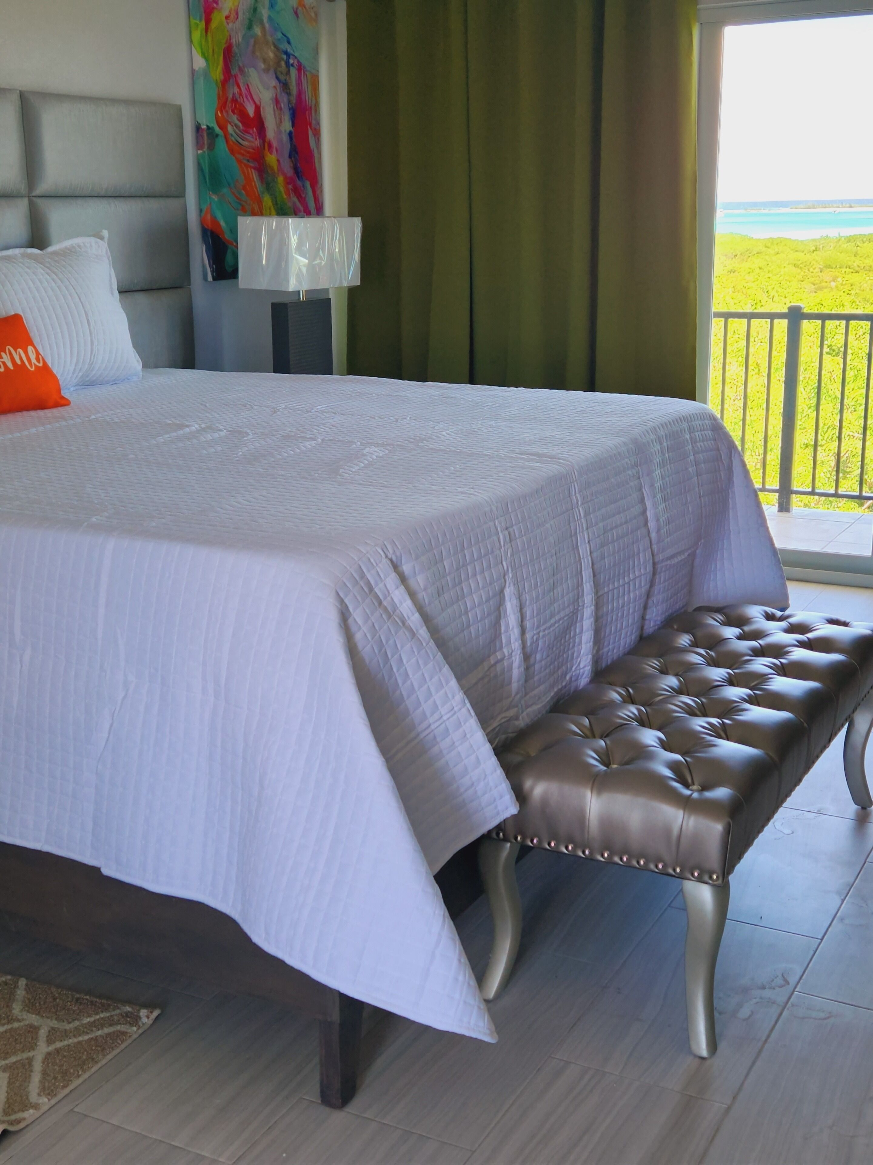 Una cama bien hecha con colcha blanca, un banco tapizado en marrón y un cuadro abstracto de colores en la pared.