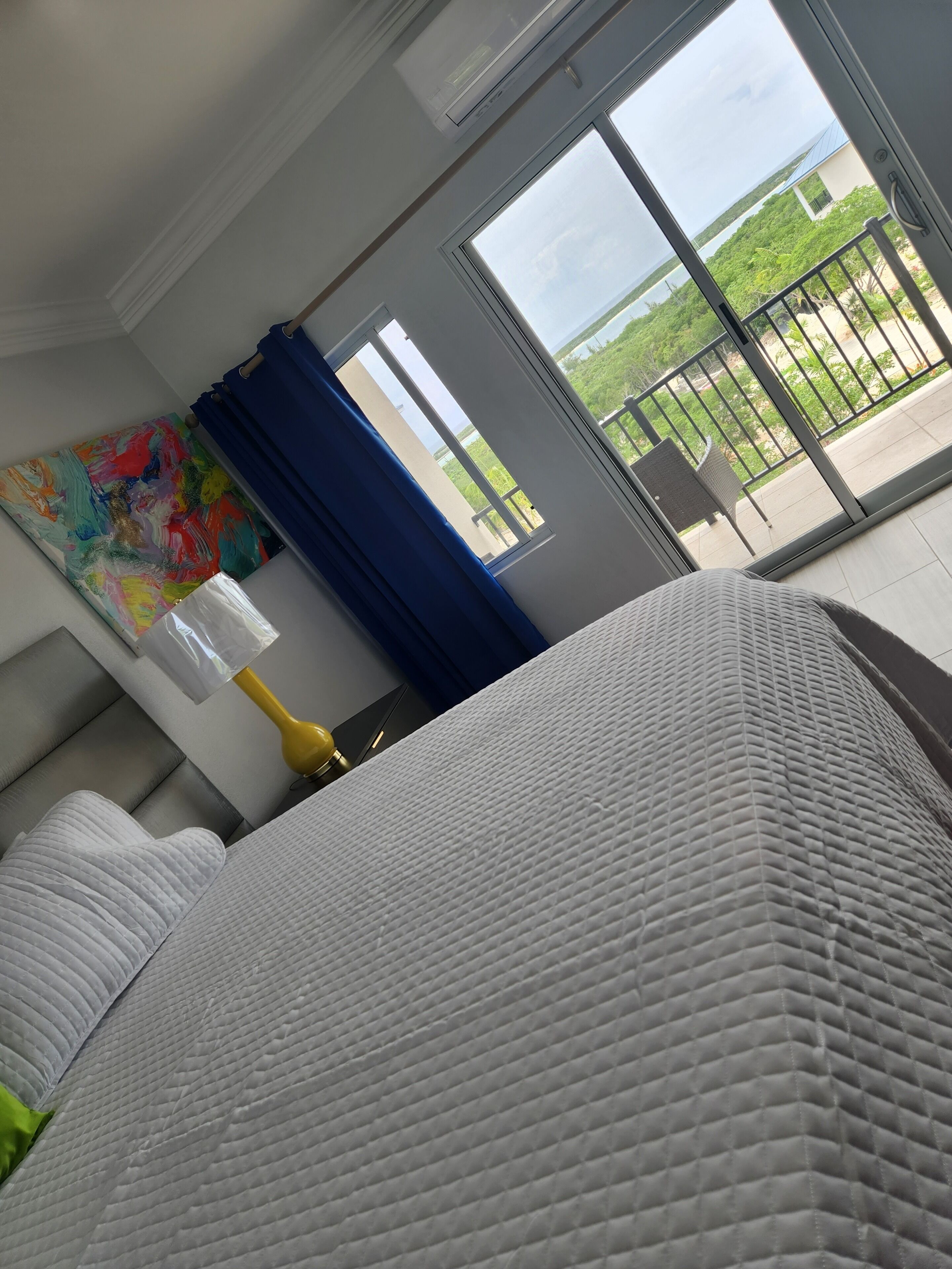 A hotel room with a bed, a painting on the wall, a window with a view of greenery, and an air conditioning unit.