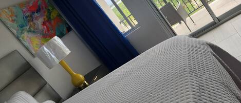A hotel room with a bed, a painting on the wall, a window with a view of greenery, and an air conditioning unit.