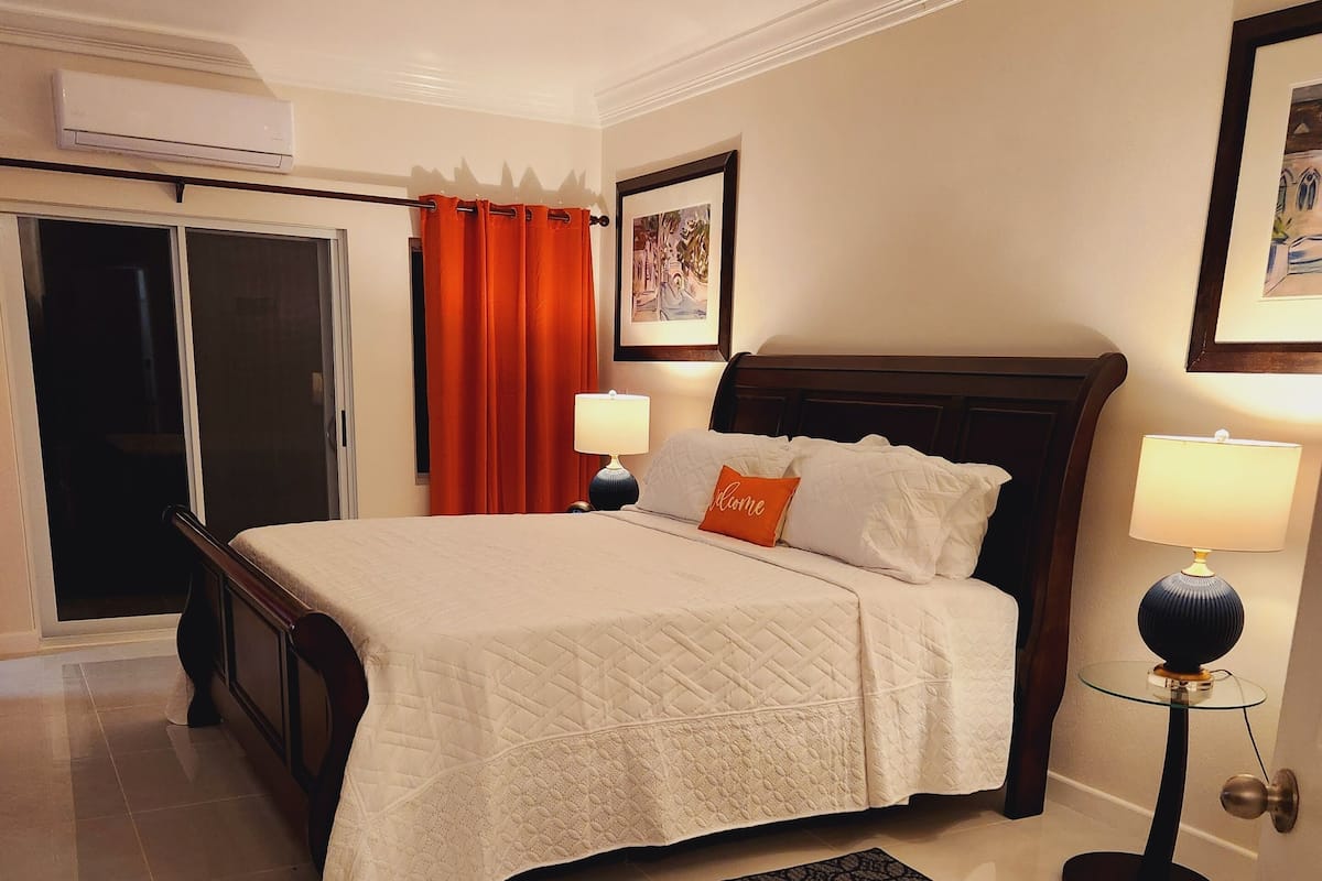 Une chambre à coucher bien rangée, avec un lit, deux lampes de chevet, un coussin décoratif, un tapis noir et un tableau encadré au mur.