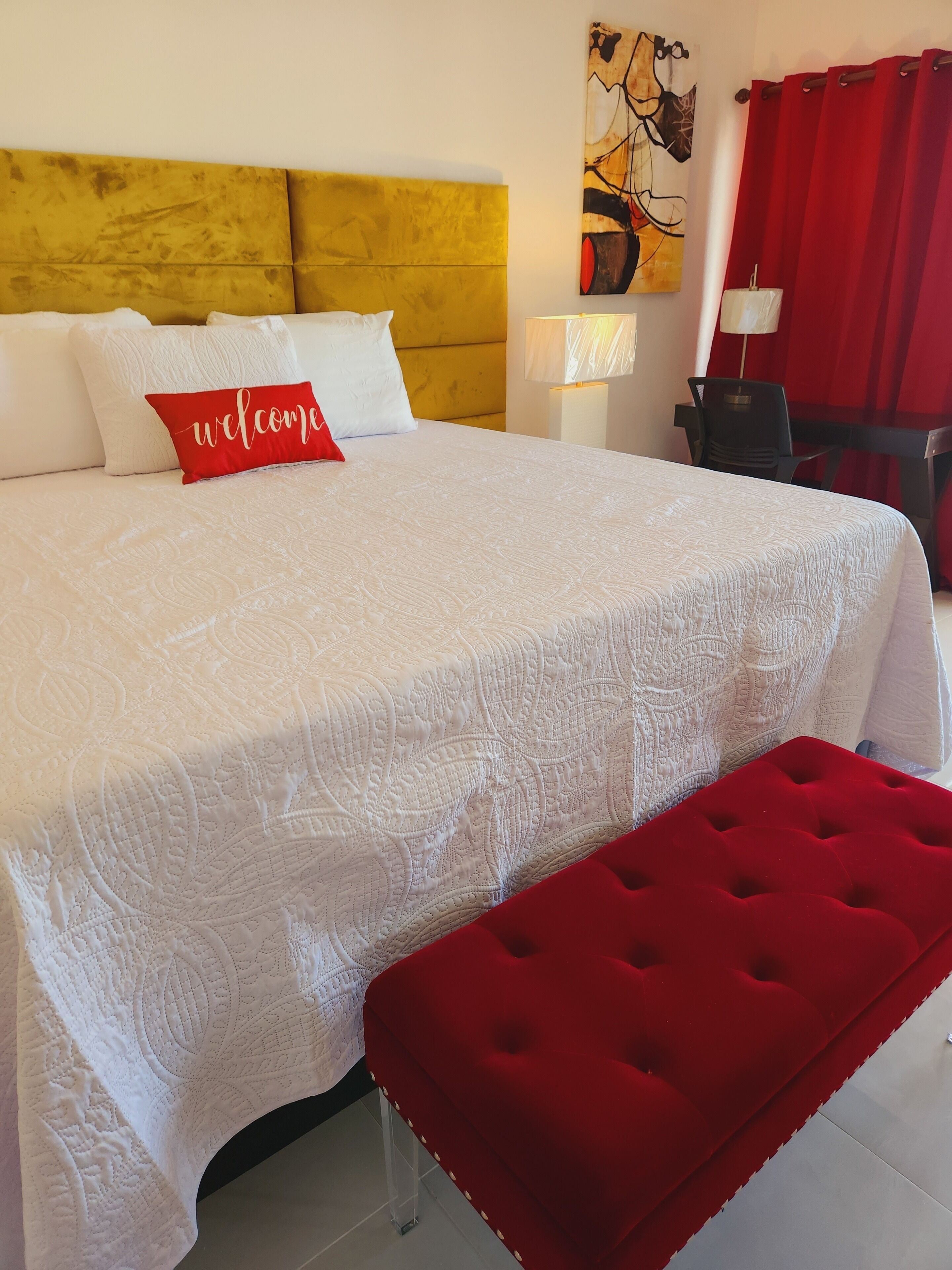Un lit bien fait, agrémenté d’un coussin rouge sur lequel est brodé le mot « bienvenue », d’un banc rouge et d’une tête de lit jaune.