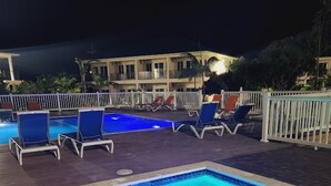Une piscine rectangulaire illuminée de lumières bleues, entourée de chaises longues et d’une clôture blanche.
