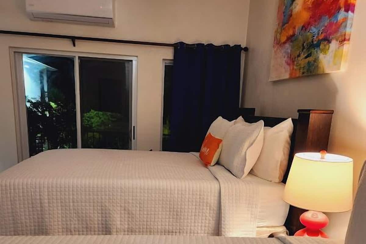 Une chambre d’hôtel avec deux lits, une fenêtre avec des rideaux, un appareil de climatisation, un tableau coloré au mur et une table de chevet avec une lampe.