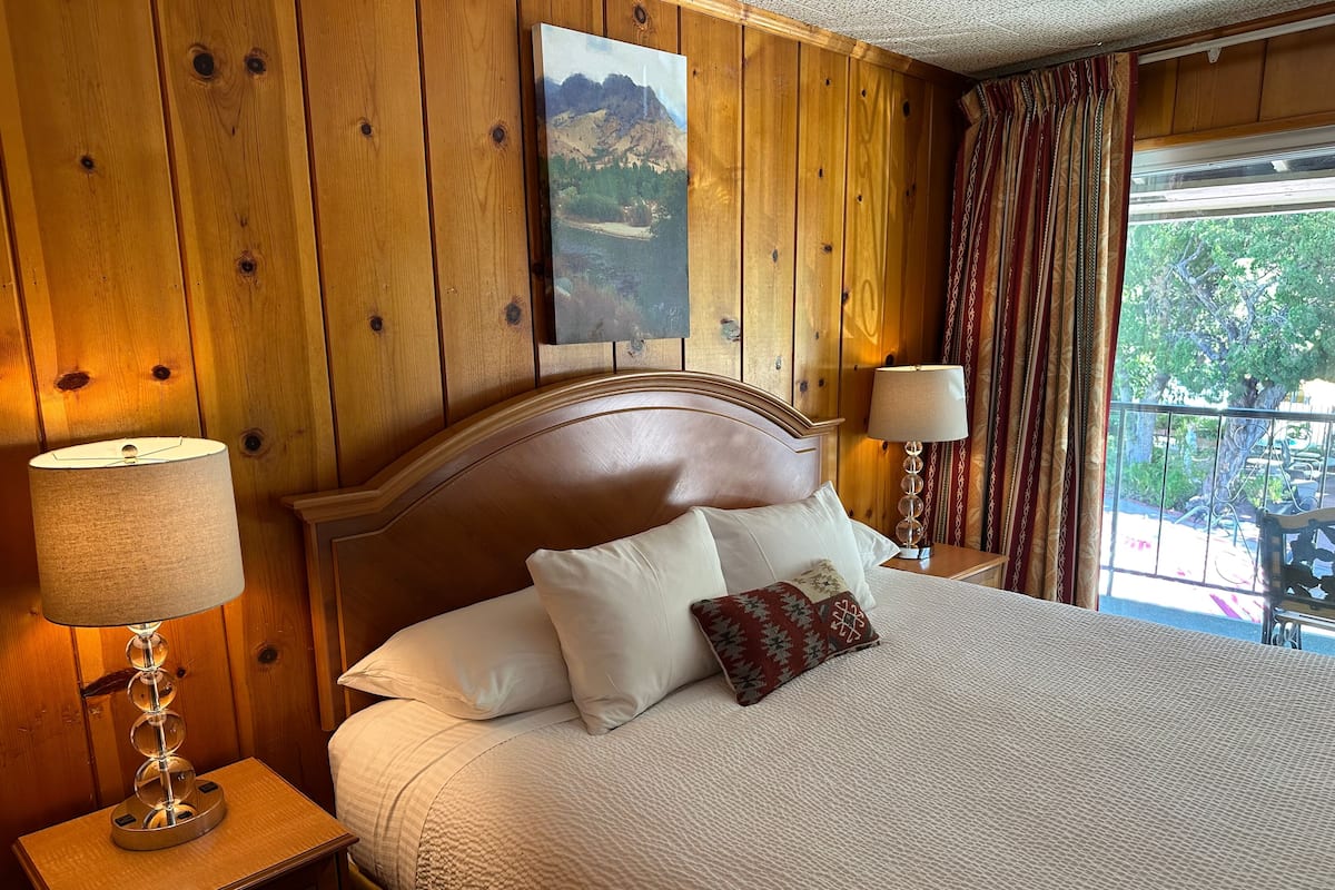 Quarto com painéis de madeira, uma cama, duas luminárias laterais e vista para o exterior.