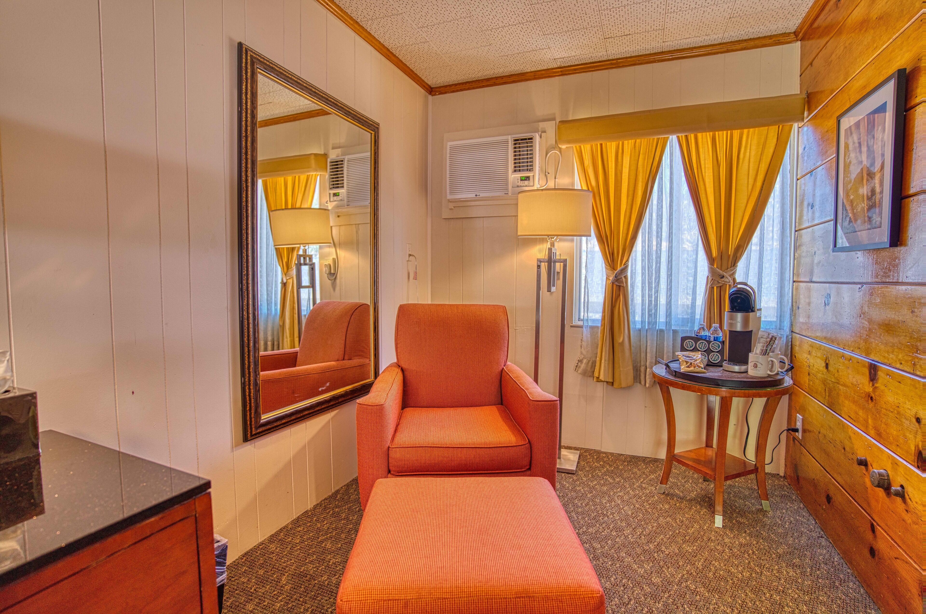 A hotel room with a wooden cabinet, a single bed, a chair, a table with a lamp, and a mirror.