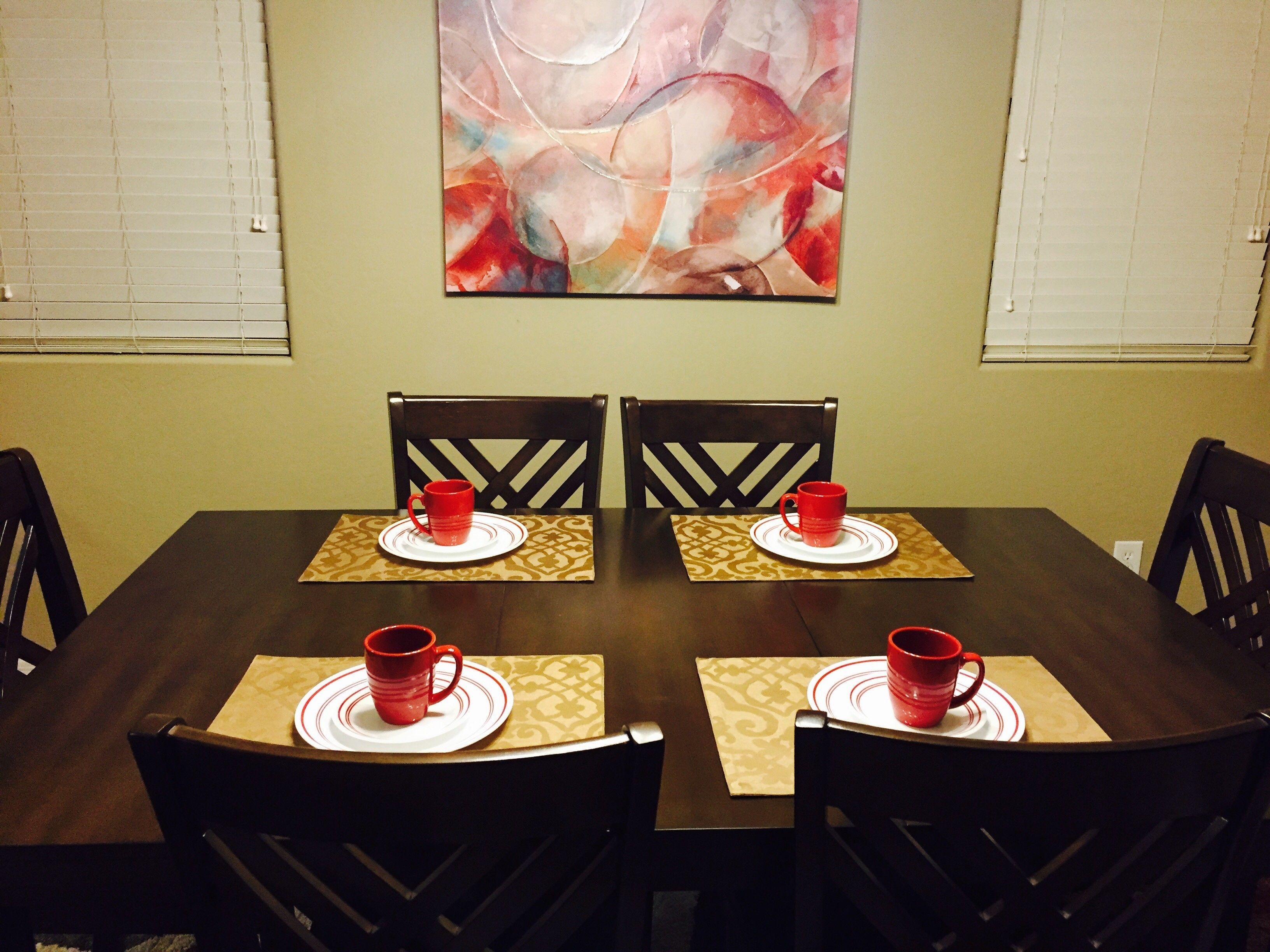 Una zona de comedor con una mesa de madera oscura, cuatro sillas y tazas rojas sobre individuales.
