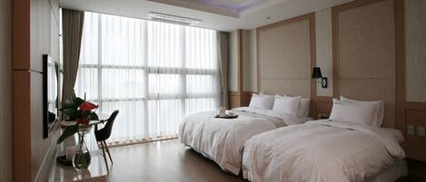 A hotel room with two beds, a desk with a chair, a large window with curtains, and a wall-mounted lamp.