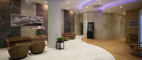 A modern lobby with a curved wooden floor, a large white pillar, wicker furniture, and a wall-mounted digital display.