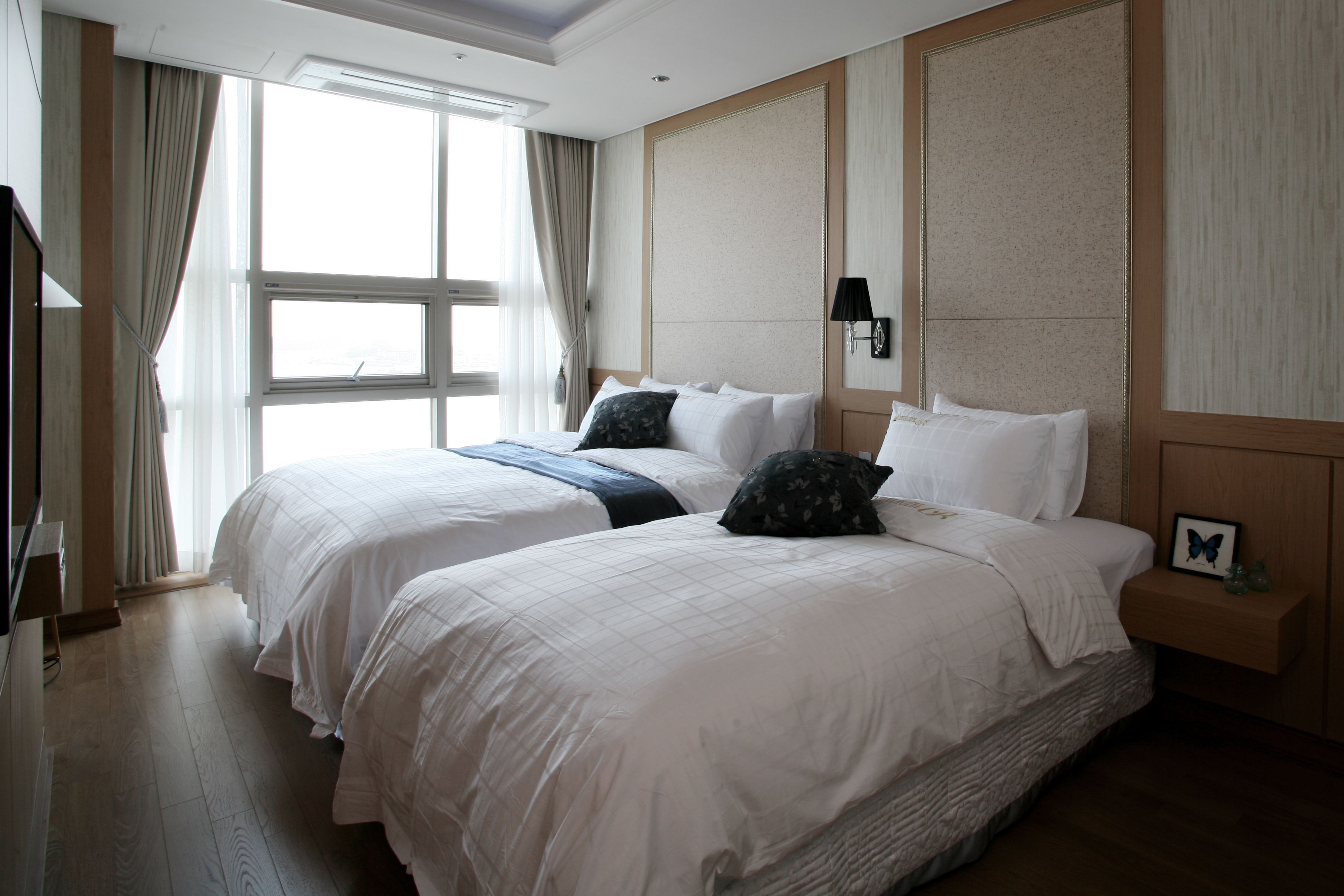 A hotel room with two beds, a large window with curtains, a wall-mounted lamp, and a small bedside table with a picture frame.
