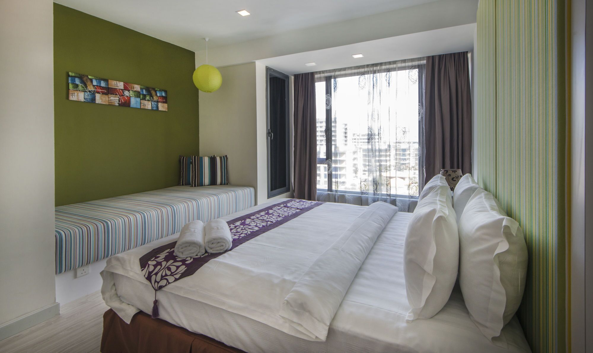 A hotel room with a large bed, a smaller bed, a window with curtains, a green wall, and a colorful artwork.