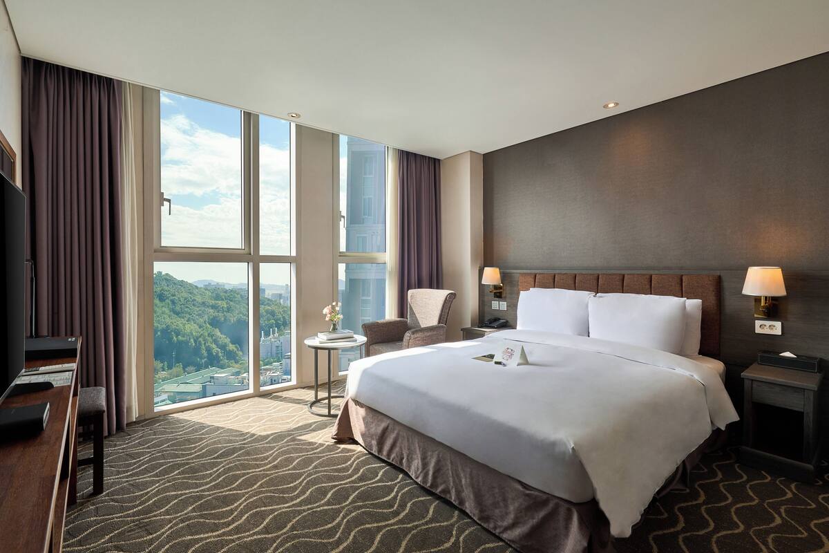 Camera d'albergo con un letto grande, una scrivania con un computer, una sedia, un tavolino con una lampada e vista sulla città attraverso ampie finestre.