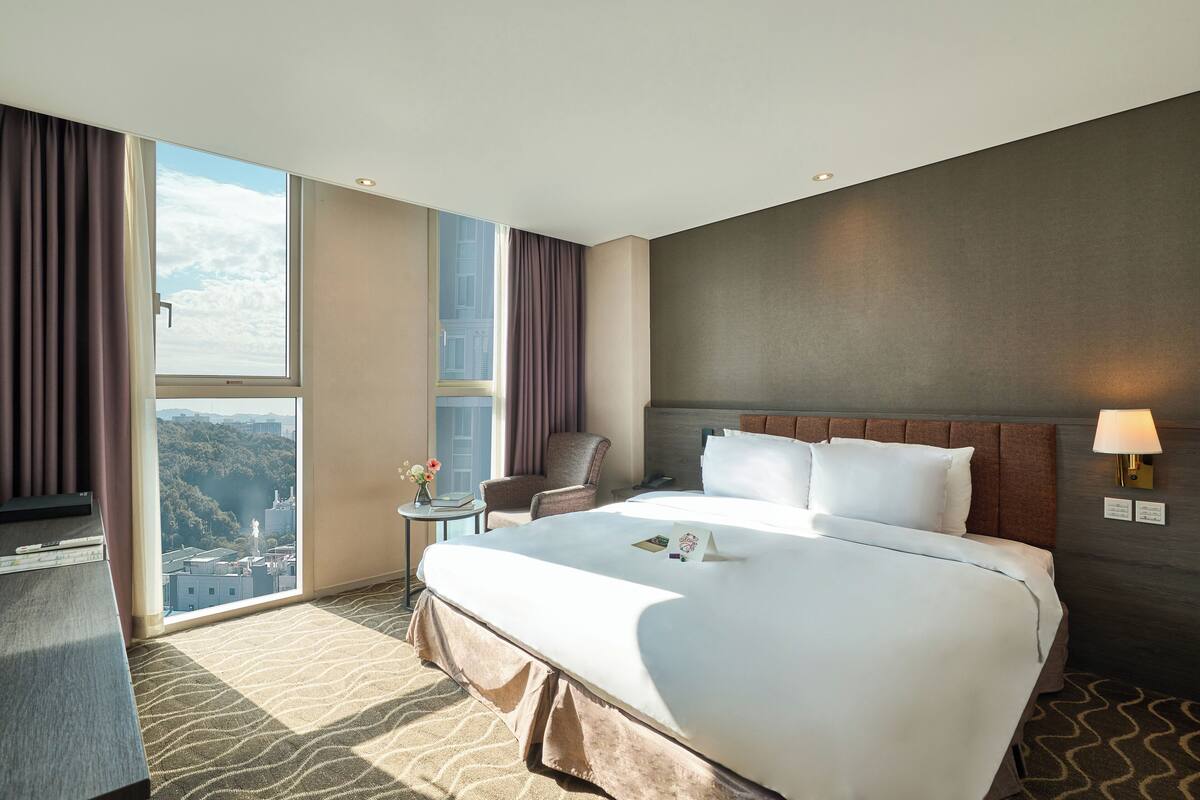 Camera d'albergo con un letto grande, una sedia, un tavolino e una finestra vista su edifici e alberi.
