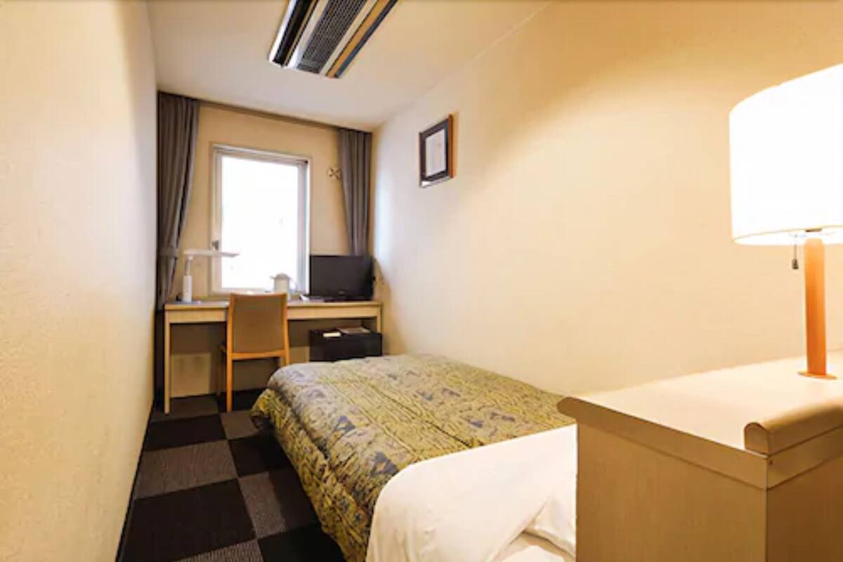 Une petite chambre d’hôtel comprenant un lit, un bureau avec une chaise, une fenêtre avec des rideaux et une lampe.
