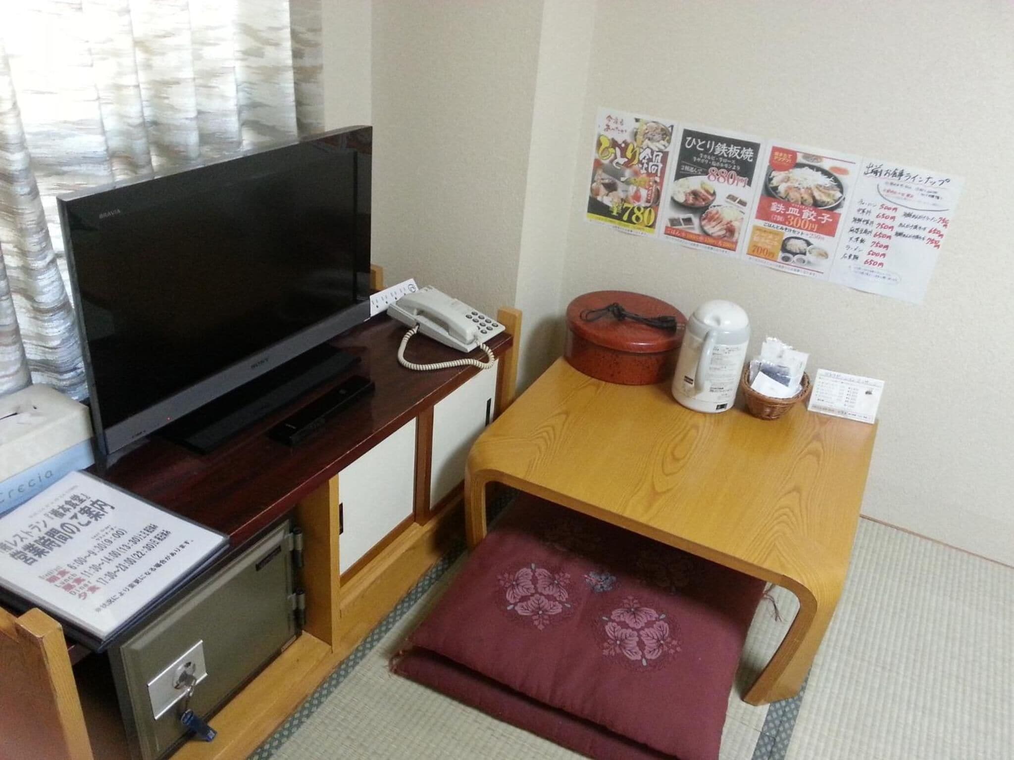 A hotel room with a television, a telephone, a small refrigerator, a wooden table with a red cushion, and a bulletin board with food menus.