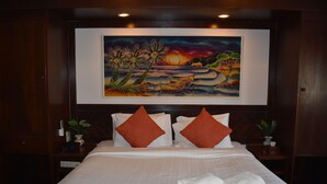 Camera d'albergo con un letto grande, biancheria da letto bianca, due cuscini arancioni, un quadro raffigurante un paesaggio al tramonto sulla parete e una piccola pianta sul comodino.