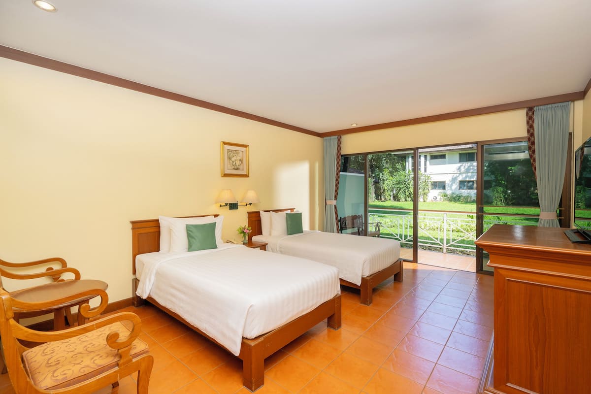 Habitación de hotel con dos camas, un escritorio, una silla y vistas a un jardín a través de amplios ventanales.