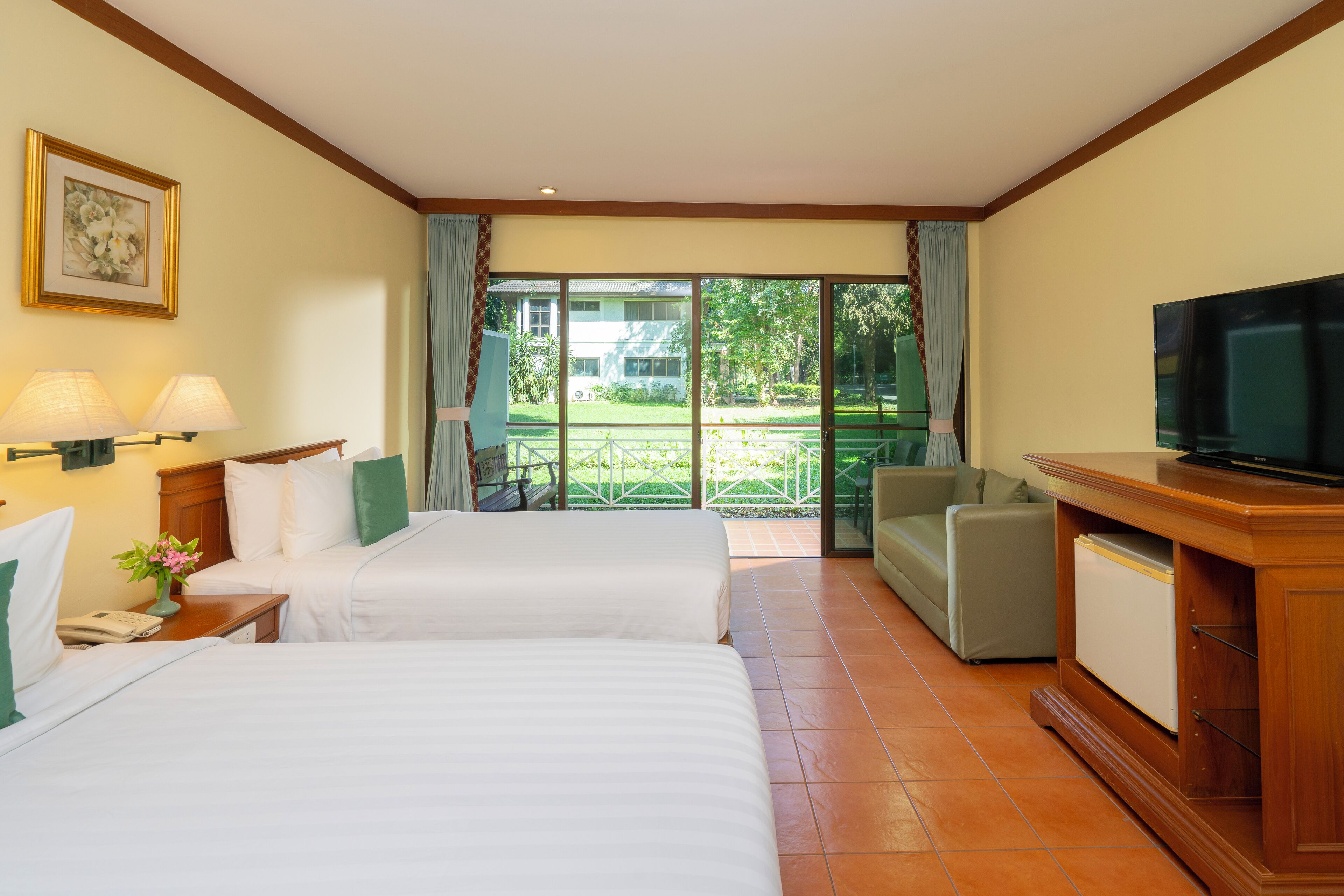 A hotel room with two beds, a television, a small refrigerator, and a view of a garden through a sliding door.