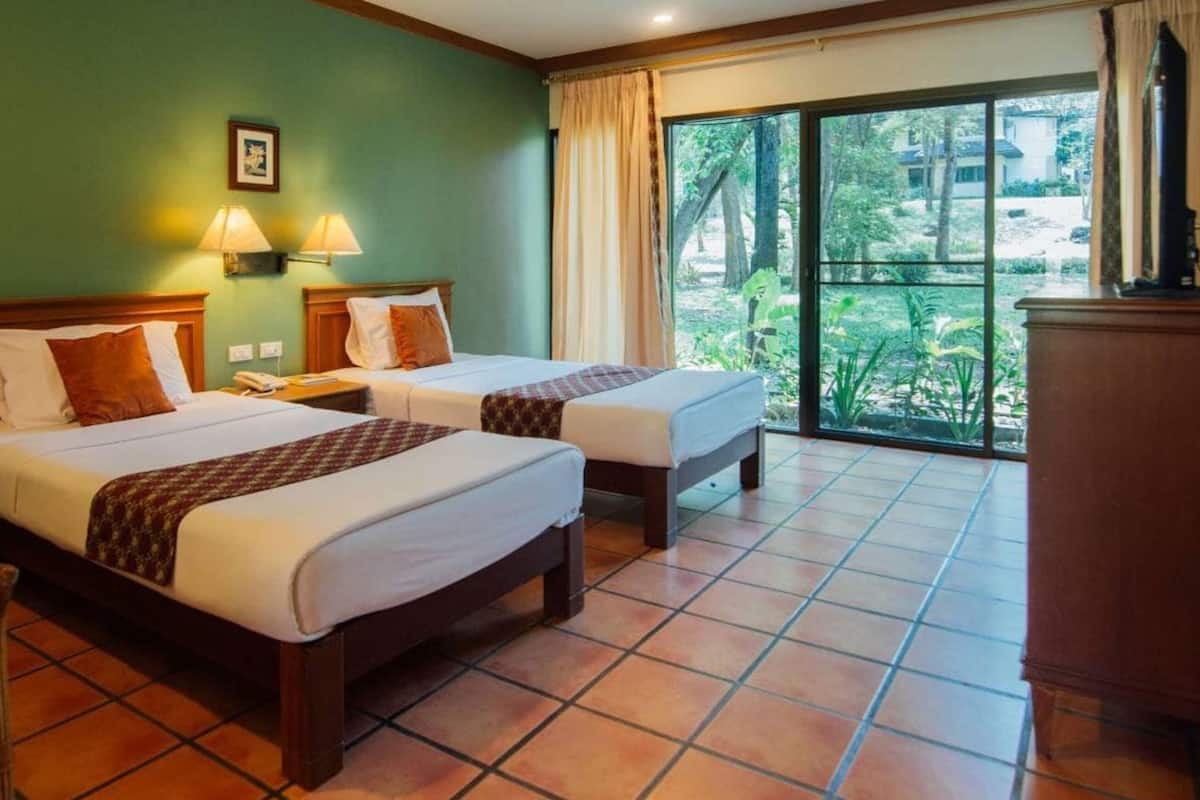 Habitación de hotel con dos camas, un escritorio, una silla, un televisor y un ventanal con vistas a zonas verdes.