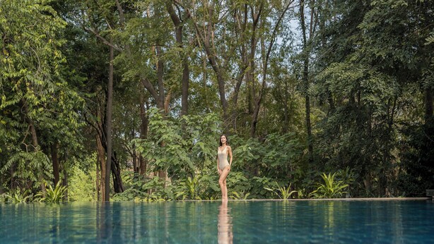 Una mujer de pie junto a una piscina cristalina rodeada de abundante vegetación.