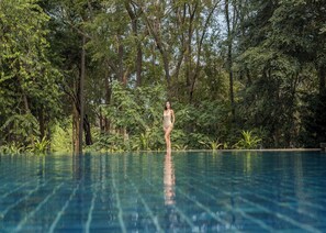 Una mujer de pie junto a una piscina cristalina rodeada de abundante vegetación.