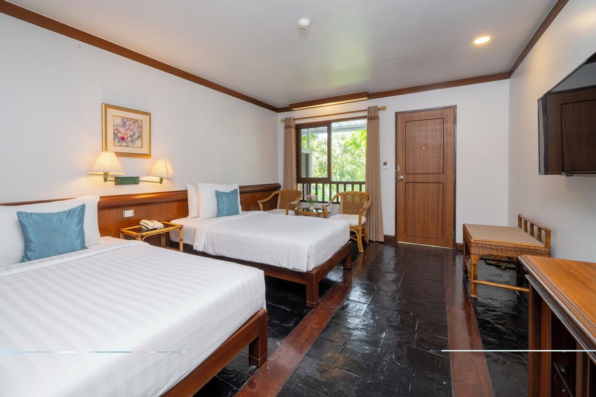 Habitación de hotel con dos camas, piso de madera, televisión y ventana con vista a follaje.