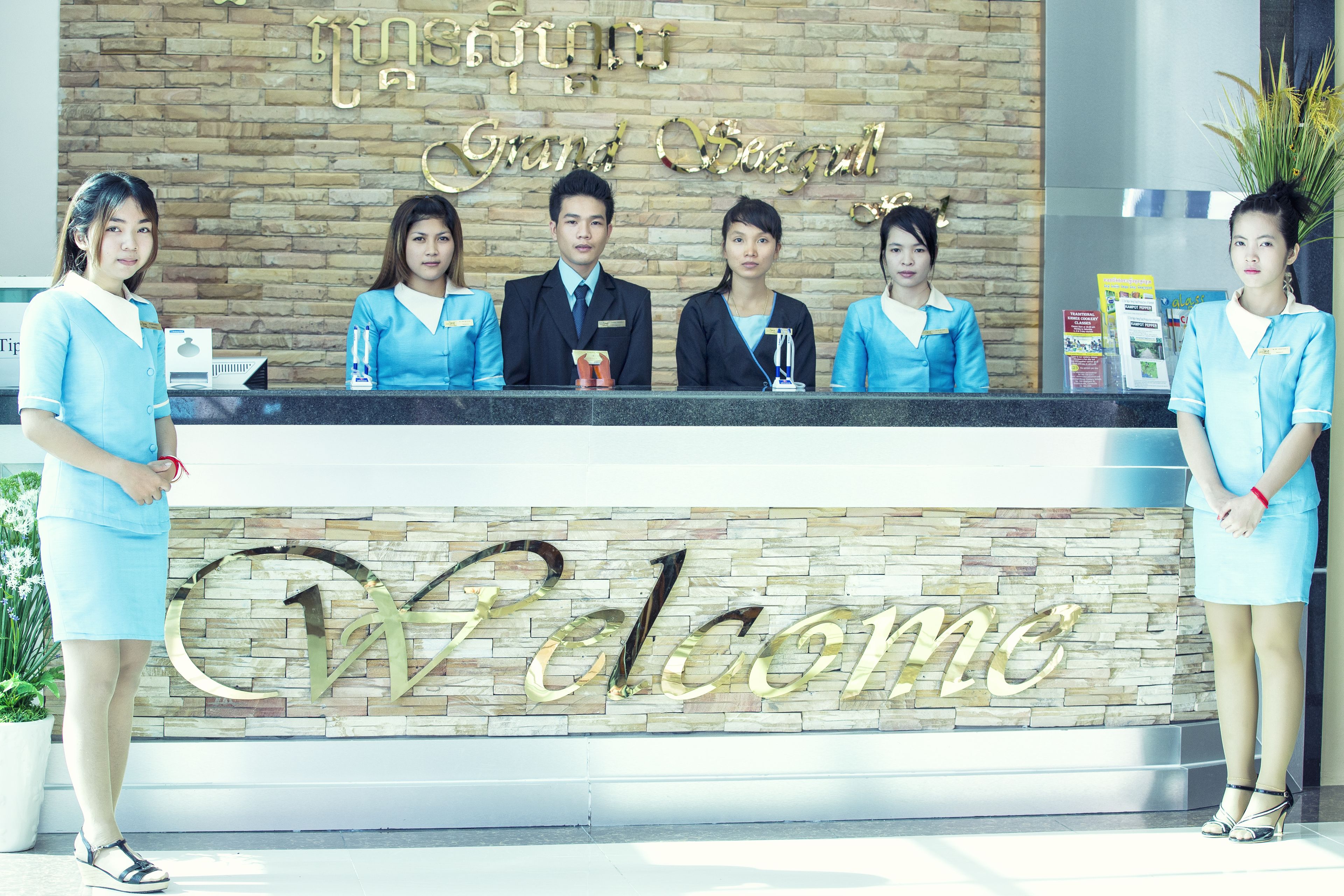 A reception desk with a "Welcome" sign, staff in uniforms, and a brick wall background.