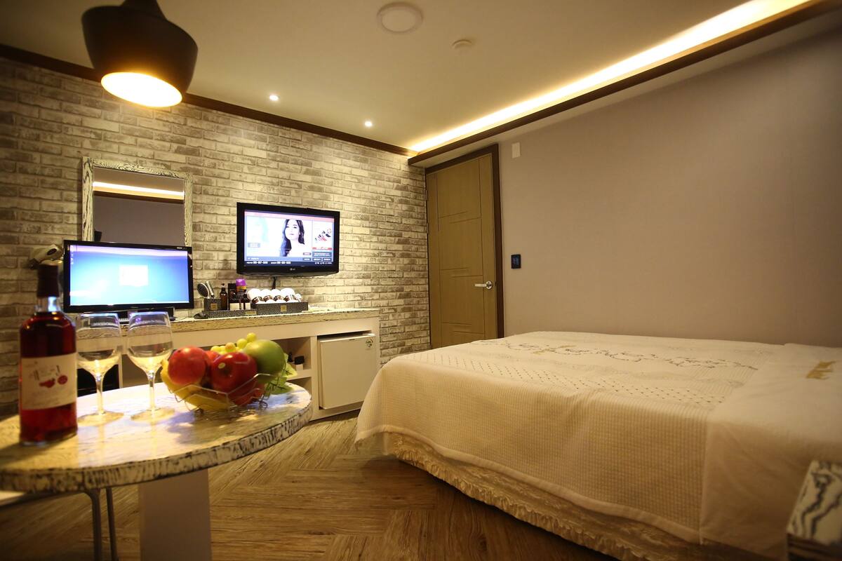 A hotel room with a bed, a TV, a desk with a computer, a wine bottle, and a fruit bowl.