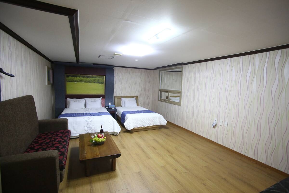 A hotel room with two beds, a sofa, a wooden table with fruit, and a view of a green field through a window.