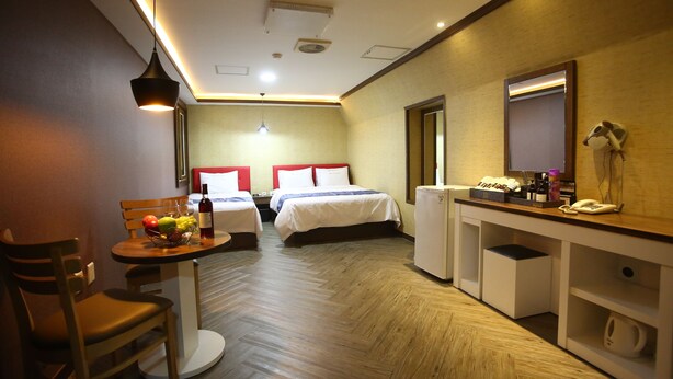 A hotel room with two beds, a small table with fruit and a bottle, a desk with a phone and a mirror, and a compact kitchenette. - Dubai Motel (Yeosu)