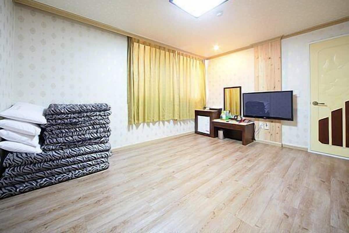 A hotel room with a wooden floor, a bed with patterned bedding, a desk with a mirror, a television, and a door.