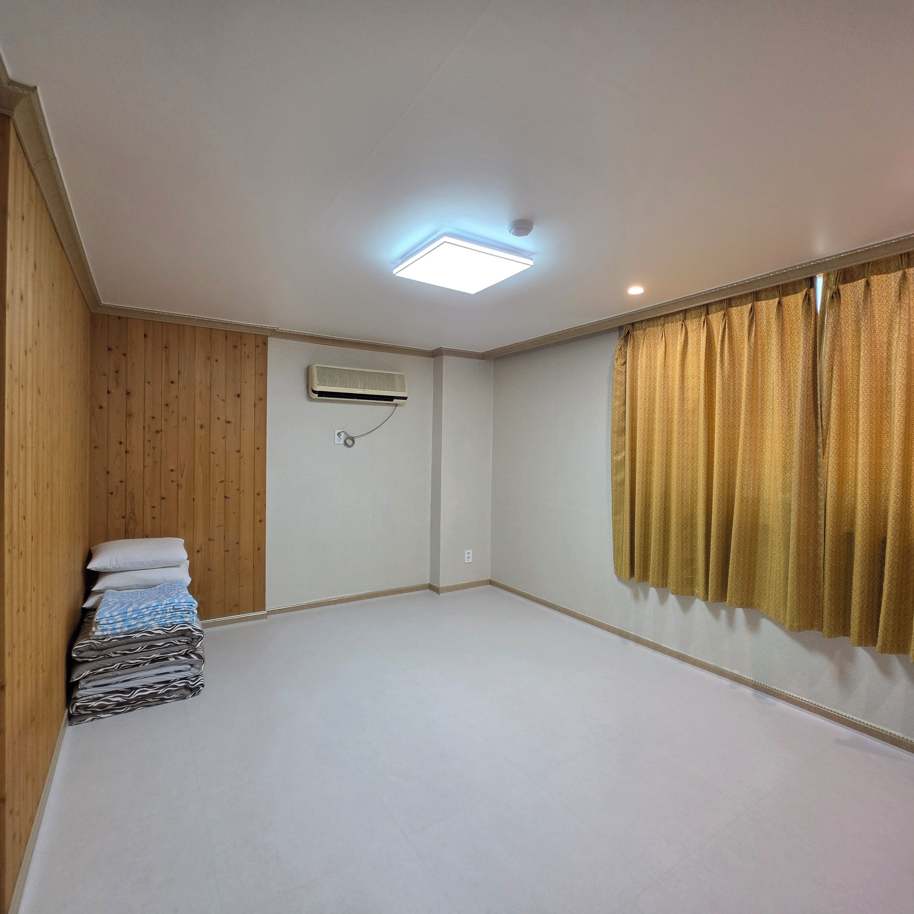 A room with a ceiling fan, an air conditioning unit, a window with curtains, and a neatly stacked pile of pillows and blankets.