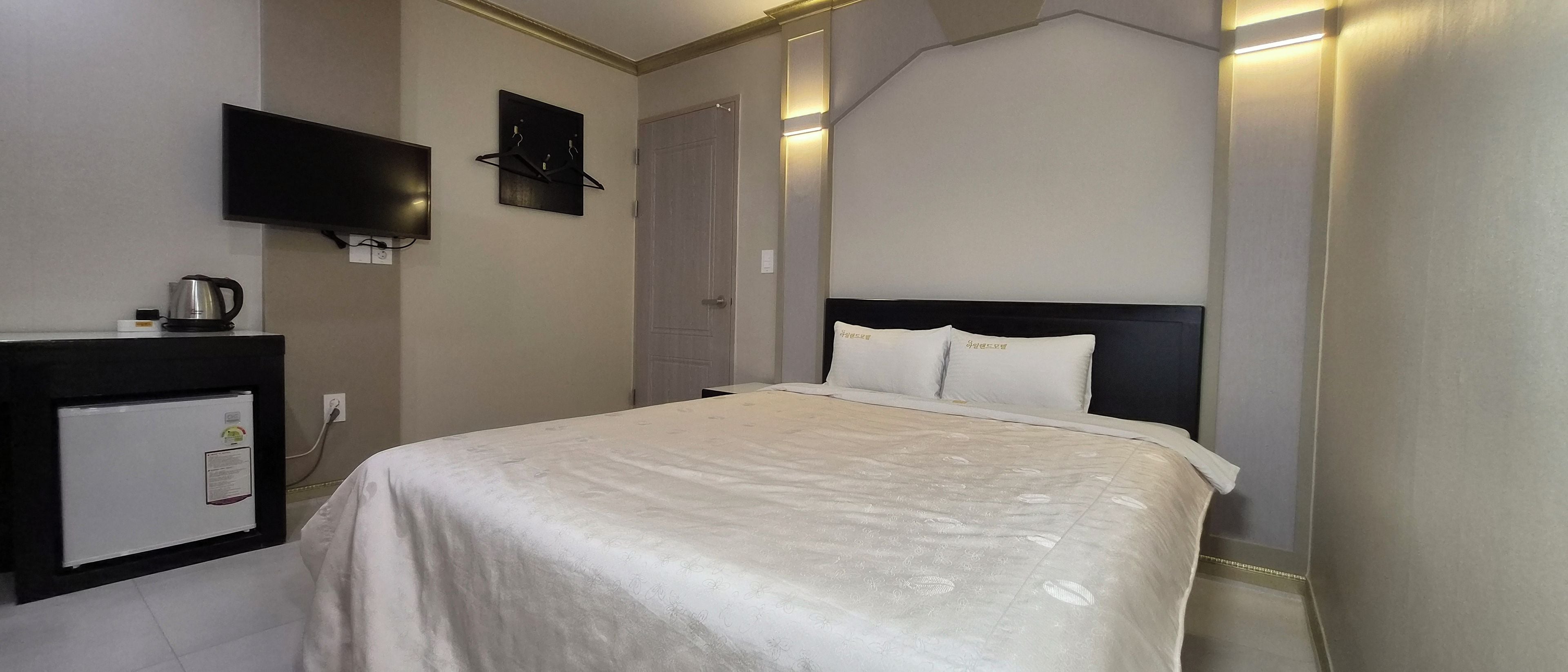 Una habitación de hotel con una cama, un televisor de pantalla plana en la pared, una pequeña zona de kitchenette con hervidor, y una puerta.