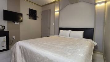 Una habitación de hotel con una cama, un televisor de pantalla plana en la pared, una pequeña zona de kitchenette con hervidor, y una puerta.