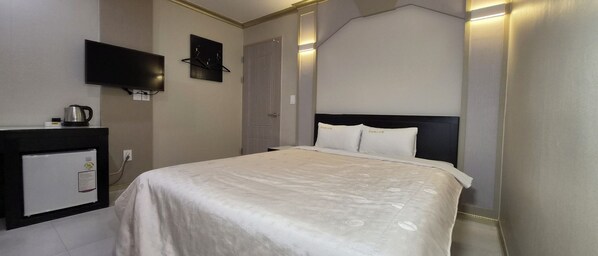 Una habitación de hotel con una cama, un televisor de pantalla plana en la pared, una pequeña zona de kitchenette con hervidor, y una puerta.