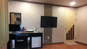 Una habitación con piso de madera, un escritorio con computadora, televisor de pantalla plana, refrigerador y una puerta con diseño decorativo.