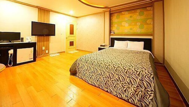 Una habitación de hotel con una cama, un escritorio con televisor de pantalla plana, suelo de madera y una puerta que da a otra habitación.