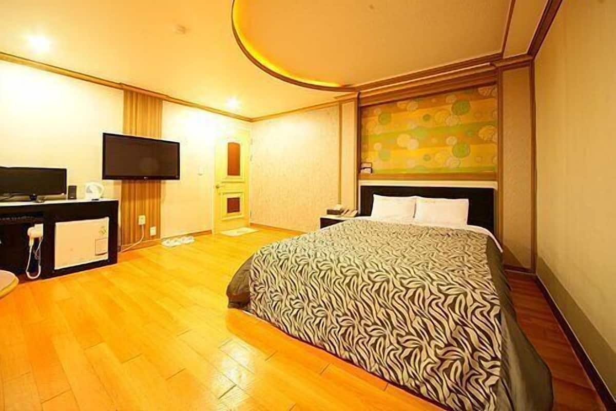 A hotel room with a bed, a desk with a flat-screen TV, a wooden floor, and a door leading to another room.
