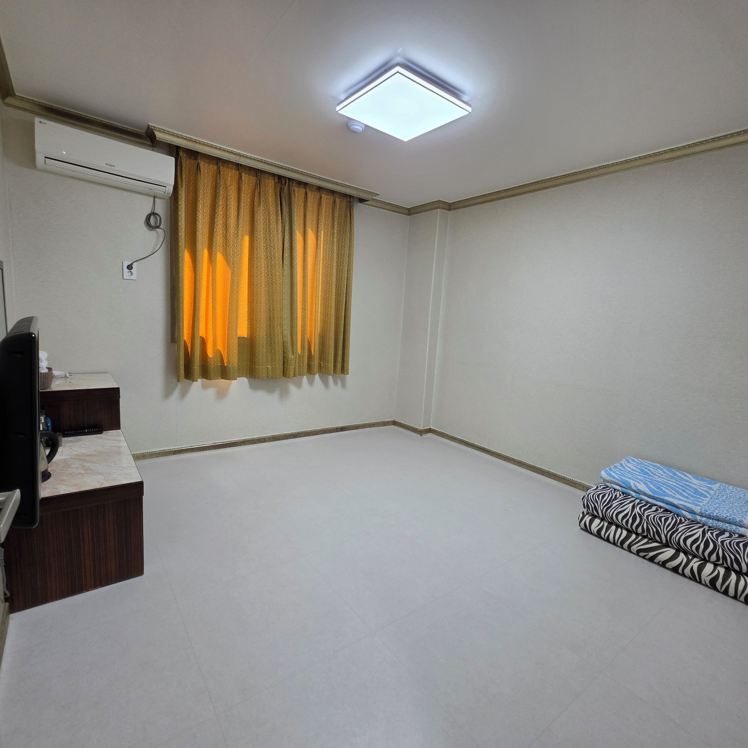 Een kamer met een plafondlamp, een airconditioning, een bureau met stoel en een bed met beddengoed met een patroon.