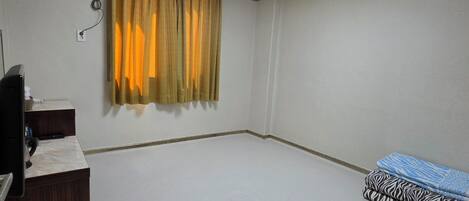 A room with a ceiling light, an air conditioning unit, a desk with a chair, and a bed with patterned bedding.