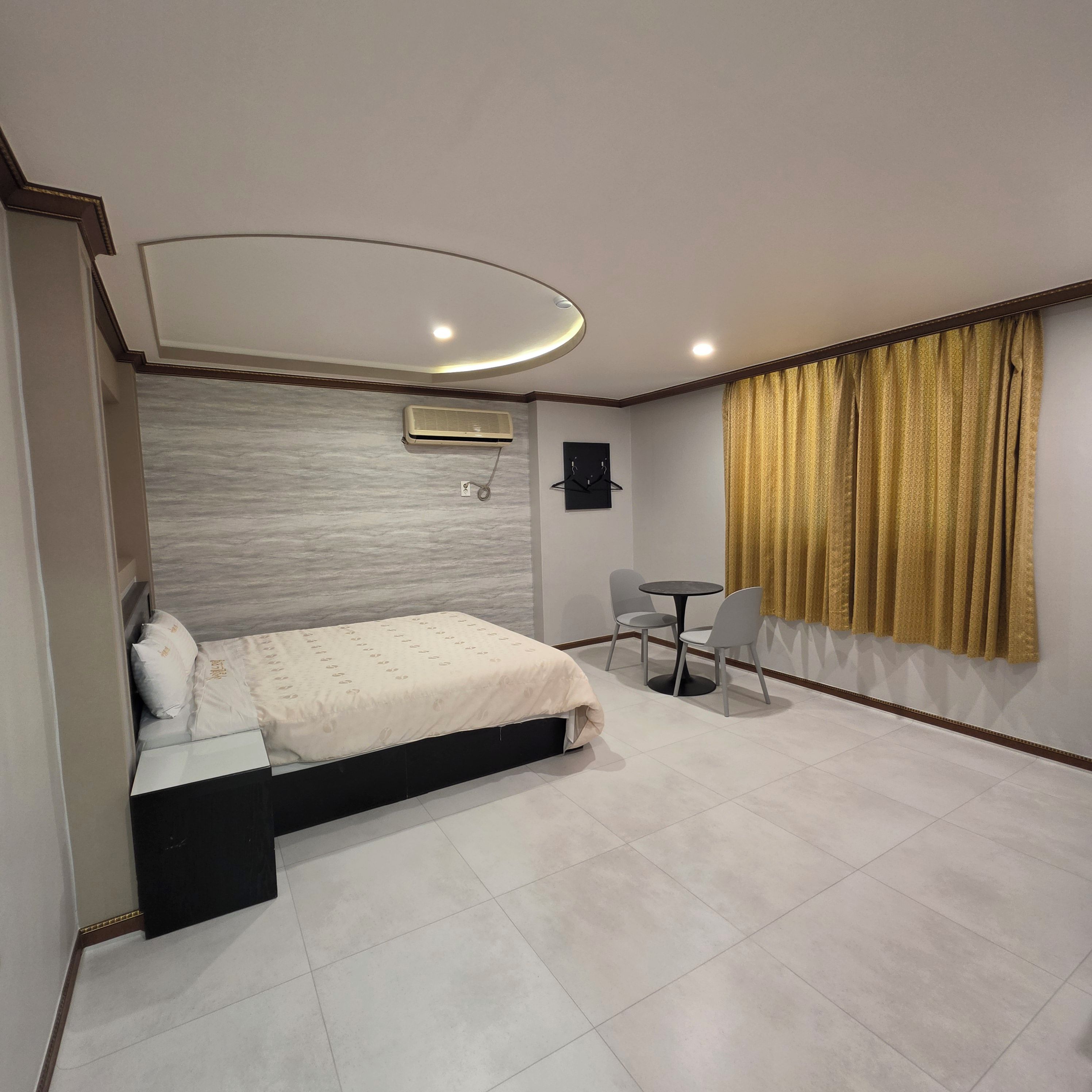 A bedroom with a bed, a small table with chairs, an air conditioning unit, and a window with curtains.