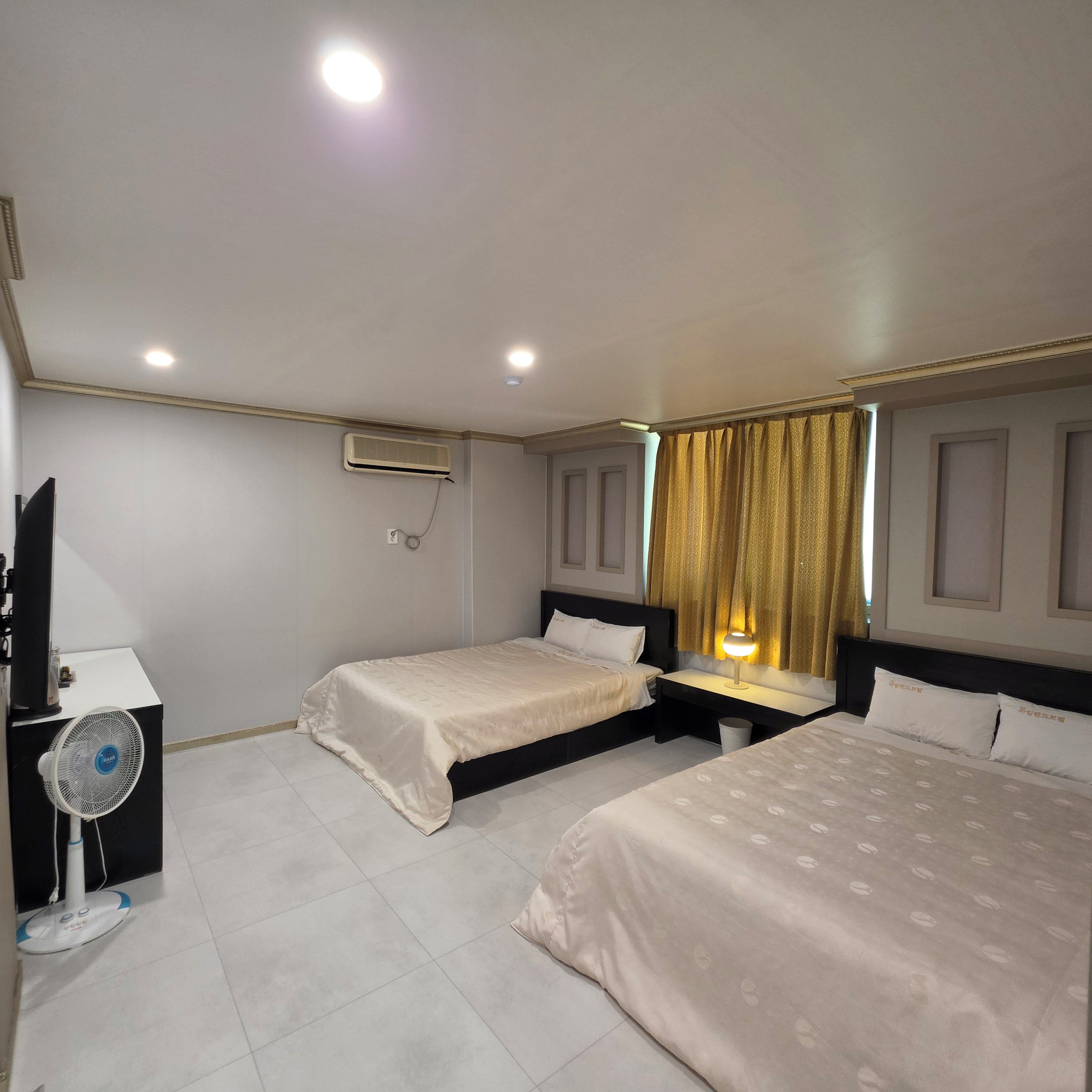 A hotel room with two beds, a desk, a chair, a TV, and a fan.