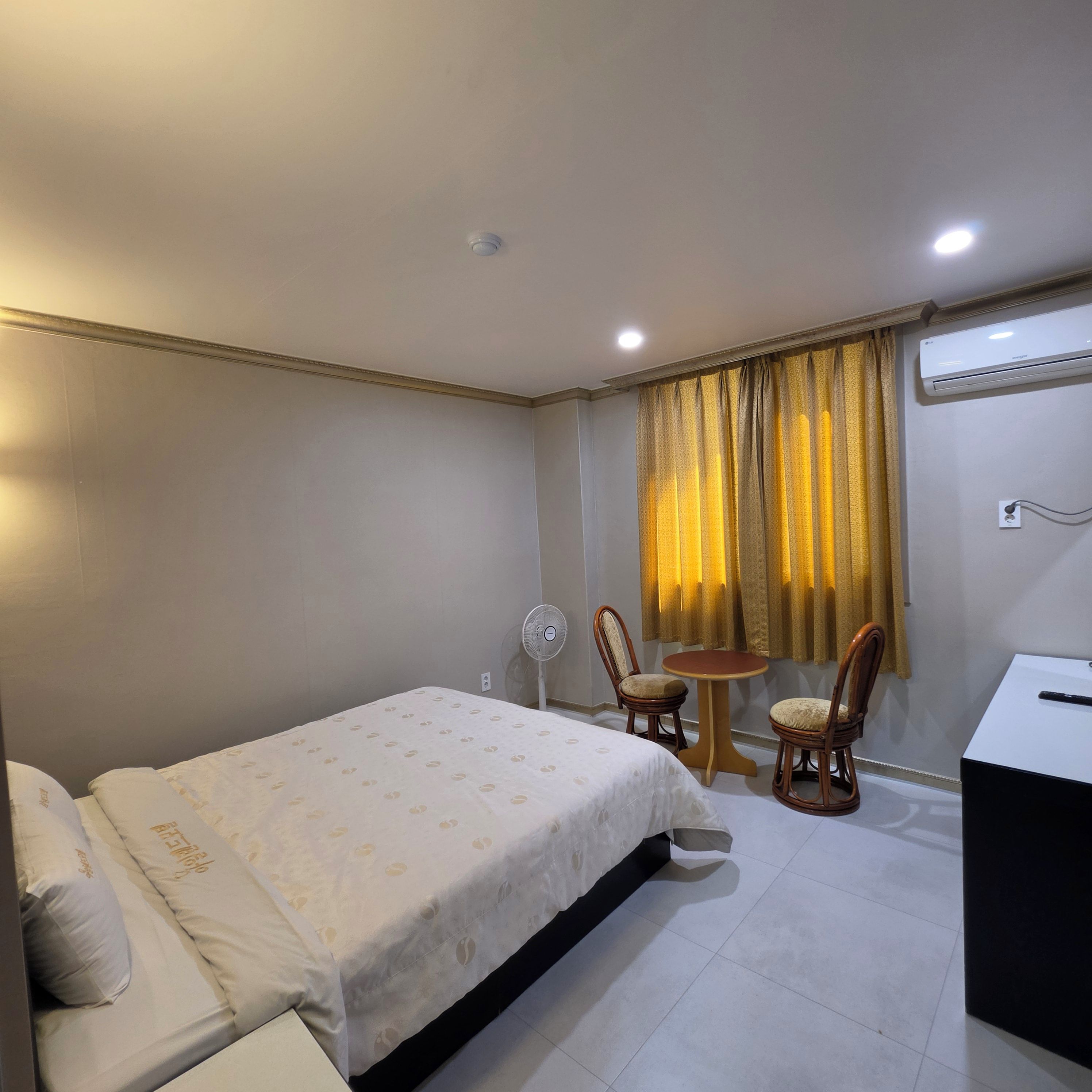 A hotel room with a bed, two chairs, a small table, an air conditioning unit, and a fan.