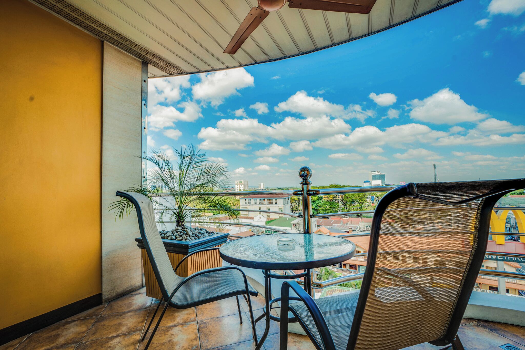 En balkon med et rundt bord og stole, en ventilator i loftet og udsigt til bygninger og skyer.