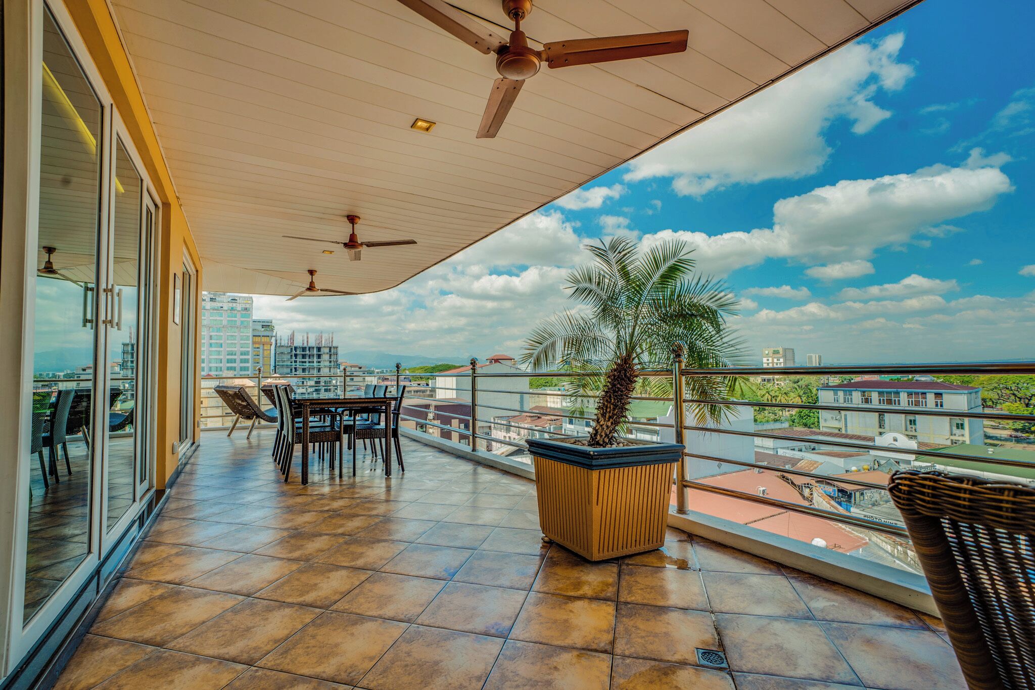 A spacious balcony with a tiled floor, ceiling fans, a dining table with chairs, and a potted plant.