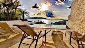 Área de piscina com espreguiçadeiras e mesa, com vista para um corpo de água e montanhas.